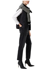 Gilet trapuntato nero sopra una camicia bianca, abbinato a jeans neri a vita alta e stivali neri con tacco a spillo. Sciarpa a righe avvolta attorno al collo.