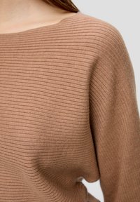 Nahaufnahme einer Person, die einen gerippten, hellbraunen Pullover mit weitem Ausschnitt trägt, wobei Details von Schulter und Oberkörper sichtbar sind.