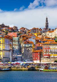 Bunte Gebäude mit Ziegeldächern entlang eines Flussufers, mit verschiedenen Baustilen, Marktständen und einem hohen Glockenturm im Hintergrund.