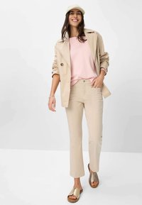 Beige trenchcoat over een roze top, licht beige broek en gouden slidesandalen. Eenvoudig ontwerp met zakken, zachte texturen en neutrale kleuren.