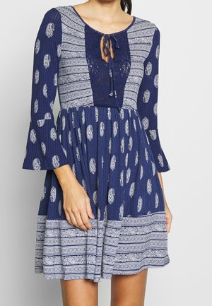Mujer con vestido azul estampado con motivos de cachemira y geométricos, mangas acampanadas y detalle de escote en V con cordones.