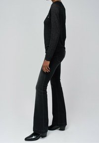 Frau in schwarzem langärmeligen Oberteil, schwarzen Schlagjeans und schwarzen Absatz-Stiefeletten, die vor einem schlichten hellgrauen Hintergrund steht.