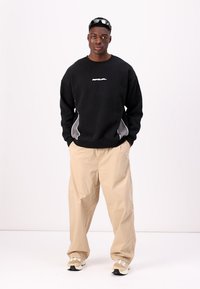Felpa nera con logo bianco, pantaloni beige a gamba larga e sneakers. Il modello sta in piedi con le braccia rilassate, indossando occhiali da sole colorati.