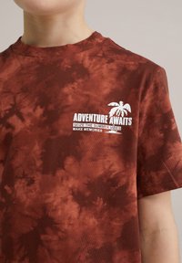 Koszulka z krótkim rękawem w brązowym tie-dye z białym napisem "PRZYGODA CZAKA" oraz grafiką palmy po lewej stronie klatki piersiowej. Wykonana z bawełny, luźny krój.