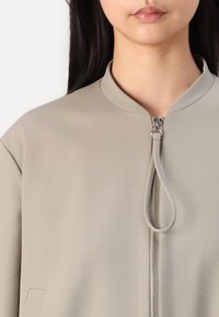 Giacca color taupe chiaro con colletto alla cinese, dotata di una cerniera frontale e un anello per la trazione. Tessuto dalla texture liscia, design minimalista.