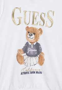 Fehér kapucnis pulóver, amelyen egy rajzolt medve látható, lila pólóban, rajta "Guess" felirat, redős szoknyával és arany fóliás betűkkel alatta.