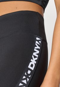 Μαύρες κολάν από ελαστικό υλικό, με λευκό λογότυπο "DKNY" κατά μήκος της πλευράς. Ο ιστός έχει λείο υφή και εφαρμοστή σχεδίαση.