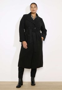 Czarny płaszcz trench z dużymi klapami, podwójnymi guzikami, paskiem w talii i bocznymi kieszeniami, w połączeniu z czarnymi spodniami i botkami za kostkę.