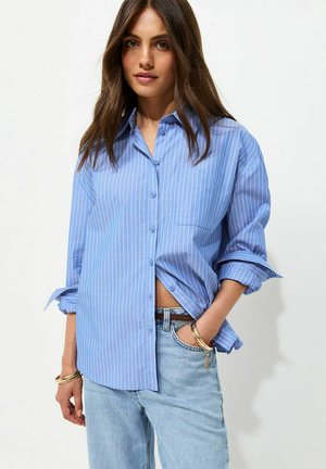 Jonge vrouw met lang bruin haar, gekleed in een los blauw gestreept overhemd en lichtblauwe spijkerbroek, met armbandjes om haar pols.