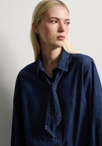 Donkerdenim overhemd met een gestrikt halsdetail, voorzien van een knoopsluiting aan de voorkant en een textuurafwerking. De kraag is gestructureerd en iets oversized.