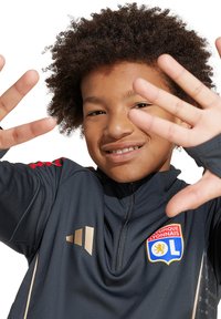 Veste de sport gris foncé avec un col haut, des accents dorés et des rayures rouges sur les épaules. Présente un logo sur la poitrine et des ouvertures pour les pouces dans les manches.