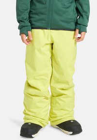 Jasné žlté lyžiarske nohavice vyrobené z vodoodpudivého materiálu. Voľný strih s pružným pásom, v kombinácii s čiernymi snehovými čižmami s bielymi podrážkami.