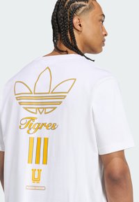 Witte katoenen t-shirt met gele grafische ontwerpen met een adidas-logo, 'Tigres' en drie strepen op de achterkant. Korte mouwen.