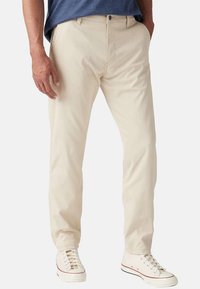 Pantalons chino en coton beige clair avec une coupe droite, dotés de poches latérales et d'une fermeture à bouton, assortis à des baskets blanches.