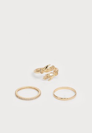 Trois bagues en or : l'une présente deux motifs de mains, l'autre a de petites pierres transparentes, et la dernière est un simple anneau poli.