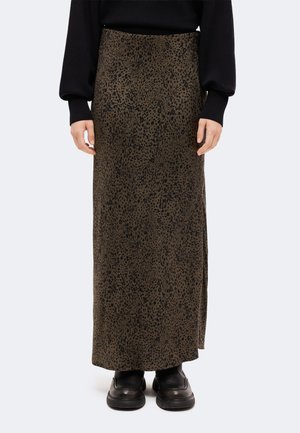 Personne portant une longue jupe à motifs marron et noir avec une fente latérale, un pull noir et des chaussures noires, debout devant un fond uni.