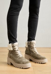 Bottines en daim vert olive avec garniture en fourrure synthétique, quincaillerie en argent et semelle plateforme beige chunky. Dispose de lacets et d'une fermeture éclair sur le côté.
