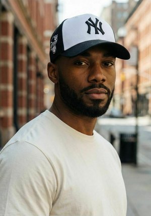 Hombre con barba que lleva una gorra de los New York Yankees en blanco y negro y una camiseta blanca, de pie en una calle de la ciudad con edificios de ladrillo al fondo.
