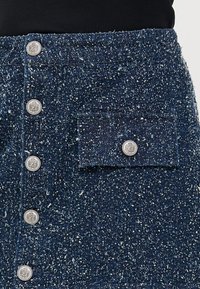 Jeanskjørt i mørkeblått med en teksturert, spraglete mønster. Har sølvknapper langs fronten og en lomme på siden.