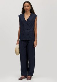 Vrouw in een mouwloos marineblauw dubbelknopsgilet en bijpassende brede broek, met een beige clutch in de hand, gekleed in kastanjebruine gestoken loafers en gouden creolen.