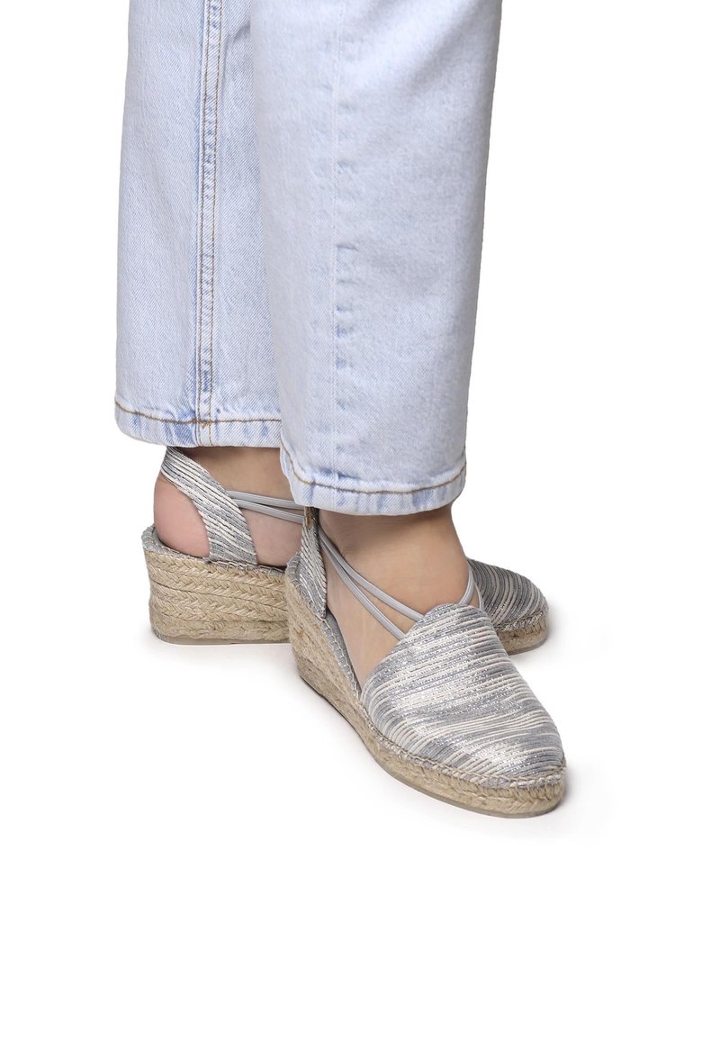 Espadrillas con zeppa e tomaia a righe argentate, materiale tessile e suola in corda di juta. Presentano cinturini incrociati alla caviglia e punta a mandorla.