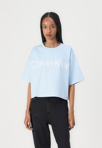 Ljusblå kortärmad t-shirt i bomull med rund halsringning, med "Calvin Klein" tryckt i vitt över framsidan, matchad med svarta jeans.