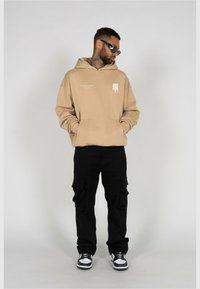 Beige hooded sweatshirt with a front graphic, paired with black cargo pants featuring side pockets and white sneakers with black accents.