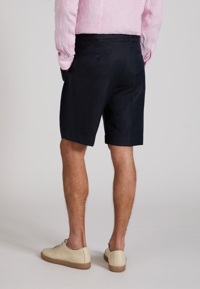 Man wearing black tailored shorts, pink long-sleeve shirt, beige sneakers, standing with hands in pockets on plain background.
