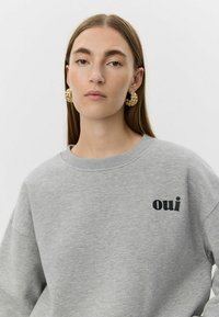 Sivá mikina s okrúhlym výstrihom a spustenými ramenami; na ľavej strane je čierny nápis „oui“. Zlaté ozdobné náušnice.