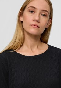 Schwarzer strukturierter Pullover mit runder Halslinie. Der Stoff hat ein dezentes geometrisches Muster, das einen dreidimensionalen Effekt erzeugt.