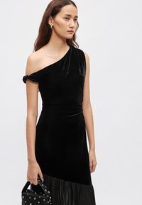 Czarna aksamitna sukienka na jedno ramię z marszczonym detalem i zwiewną jedwabną spódnicą. Modelka trzyma małą czarną torebkę o fakturowanej powierzchni.