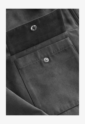 Tela de ante gris con una textura suave que presenta un cierre de botón a presión y un bolsillo. El detalle de cremallera a lo largo del borde añade funcionalidad.