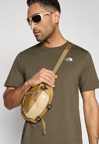 Ταν τσάντα ώμου με ρυθμιζόμενο λουράκι, μπροστινή τσέπη με φερμουάρ, μαύρα κλιπ και λογότυπο. Πράσινο ελιάς μπλουζάκι. Γκρι φόντο.