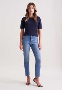 Granatowy sweter dziany z krótkimi rękawami, w połączeniu z jasnoniebieskimi dopasowanymi dżinsami i czarnymi sandałami na obcasie. Prosta, smukła sylwetka.