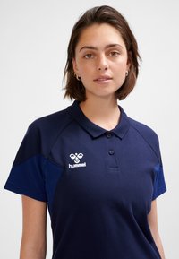 Marinblå pikétröja med tvåfärgad design, utrustad med krage, knapplista och en broderad logotyp på bröstet. Tillverkad av bomullsblandning.