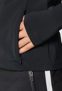 Jachetă neagră cu fermoar, având o textură netedă, dotată cu un buzunar lateral și o croială ajustată. Manșetele au un finisaj curat, bine definit.