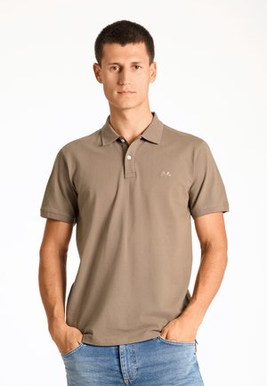 Polo marrone in cotone, con patta a tre bottoni, maniche corte e un piccolo logo sul lato sinistro del petto. Indossato con jeans blu.
