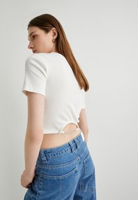 Top corto blanco de canalé con mangas cortas, que presenta un detalle de corte en la parte posterior y un acento de cadena plateada. Combinado con jeans azul de tiro alto.