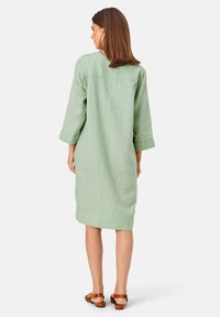 Jasnozielona lniana sukienka z rękawami o długości trzech kwart, prostym kroju i prostym wzorem z tyłu. Noszona z brązowymi sandałami na paski.