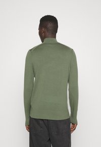 Maglione leggero di colore verde scuro, con colletto alto e maniche lunghe. Presenta una vita aderente e una finitura testurizzata sulle cuciture delle spalle.