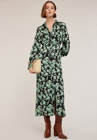 Rochie neagră cu mânecă lungă, cu un model floral verde, guler, fante laterale și croială ajustată, asortată cu cizme maro cu toc și o geantă bej.