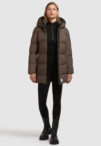 Braune Daunenjacke mit Kapuze, mit Reißverschluss vorne, gestepptem Design und Seitentaschen. Getragen über einem schwarzen Oberteil und kombiniert mit schwarzen Hosen und Stiefeln.