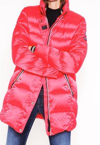 Veste rembourrée d'un rouge vif avec un col haut, une fermeture éclair complète et des zips sur les côtés. Présente des rayures noires contrastantes sur les manches et l'ourlet.