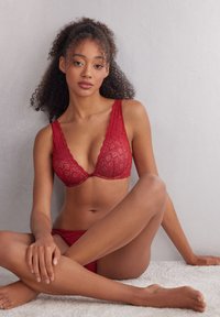 Roter Spitzen-BH mit tiefem Ausschnitt und gewellten Kanten, der zarte Diamantmuster aufweist; passende Spitzenstrings sind ebenfalls sichtbar.