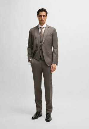 Costume trois pièces gris comprenant une veste ajustée, un gilet avec trois boutons et un pantalon fuselé. Associé à une cravate claire et des chaussures noires.