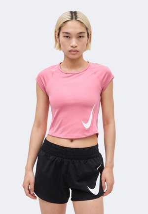 Jeune femme aux cheveux blonds courts portant un T-shirt court rose Nike et un short de sport noir Nike, debout devant un fond clair.