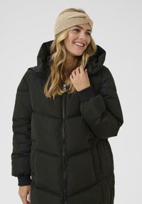 Cappotto puffo nero con zip frontale, colletto alto e tasche laterali. Indossato con una fascia per capelli beige, caratterizzata da un design a torsione e una texture morbida.