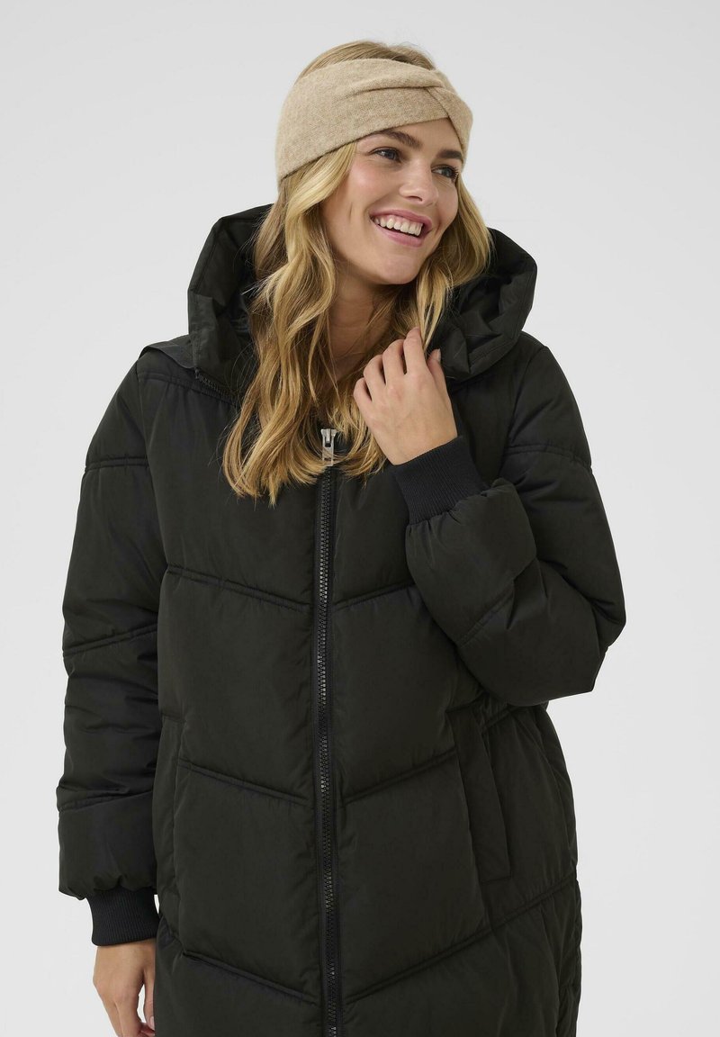 Cappotto puffo nero con zip frontale, colletto alto e tasche laterali. Indossato con una fascia per capelli beige, caratterizzata da un design a torsione e una texture morbida.