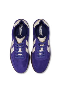 Fioletowe zamszowe sneakersy z białymi akcentami w kształcie zygzaka, płaskimi sznurowadłami i brązową gumową podeszwą. Wnętrze wyposażone w niebieską wkładkę z logo.