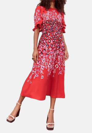 Vrouw draagt een rode midi-jurk met roze bloemen- en luipaardprint, met gerimpelde korte mouwen, gecombineerd met beige sandalen met hoge hakken.